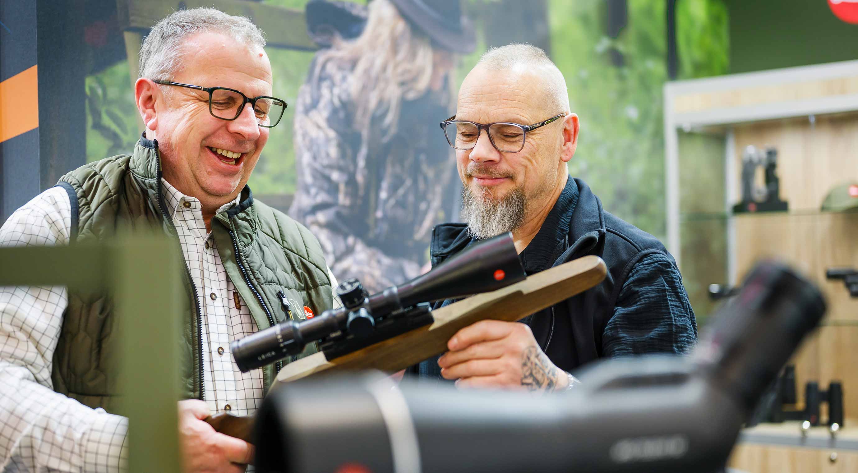Two people are looking at a rifle with a scope at a stand for outdoor and hunting gear.