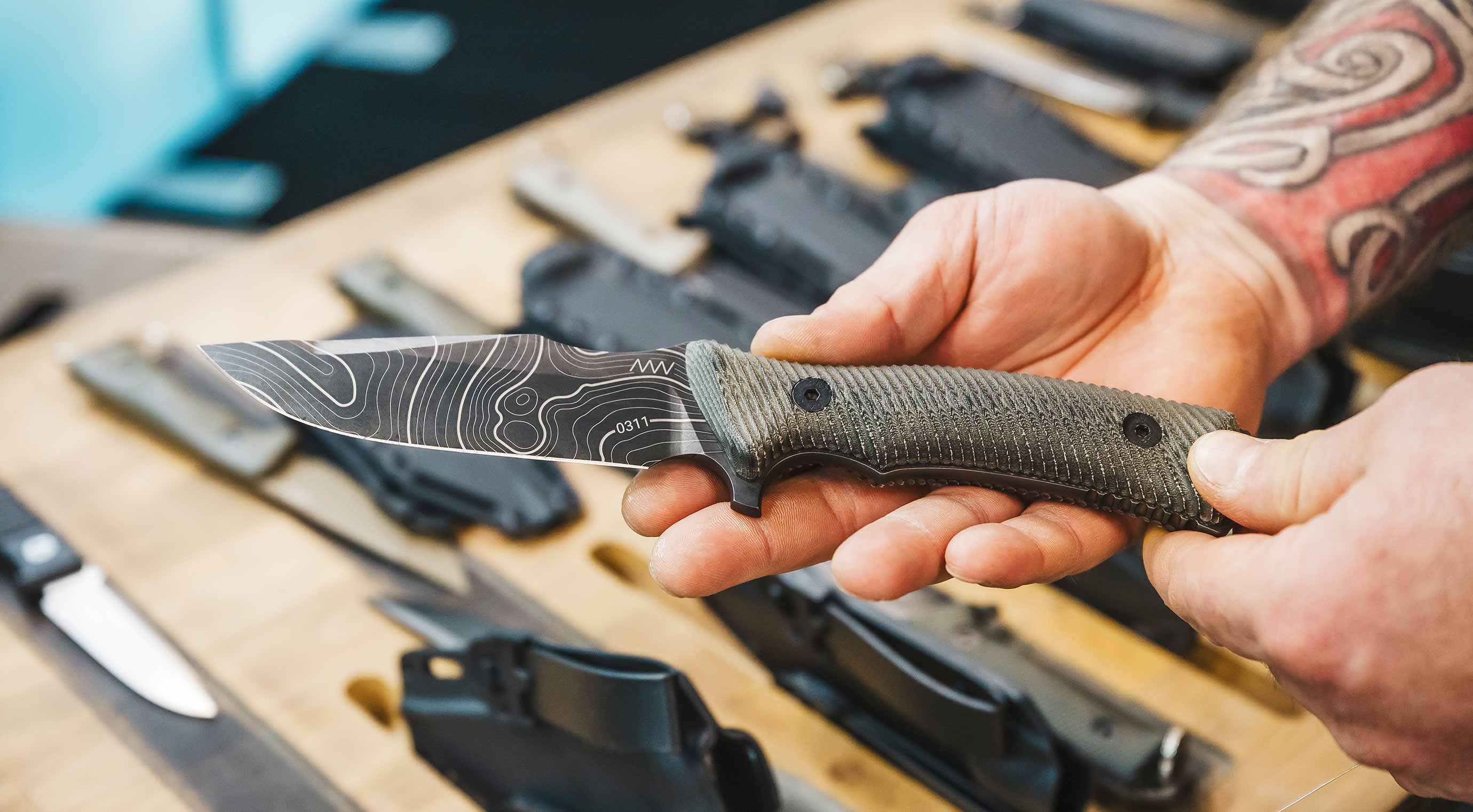 A hand holds a fixed-blade knife with a textured blade and handle, surrounded by other equipment.