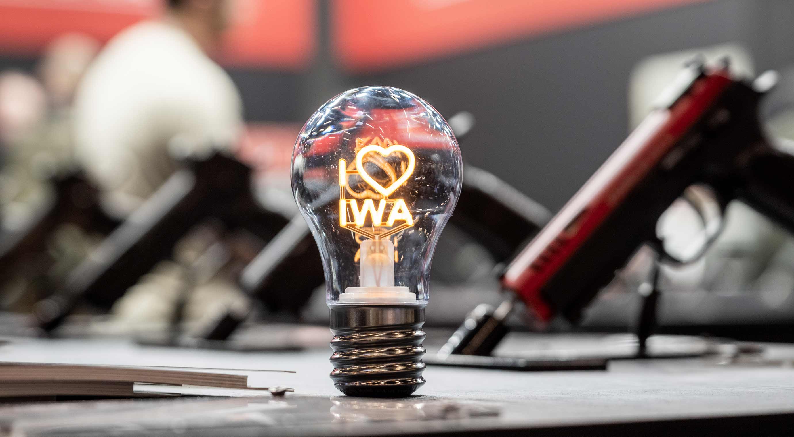 A light bulb with the illuminated lettering “I love IWA” stands on a trade fair counter. Blurred weapons can be seen in the background.