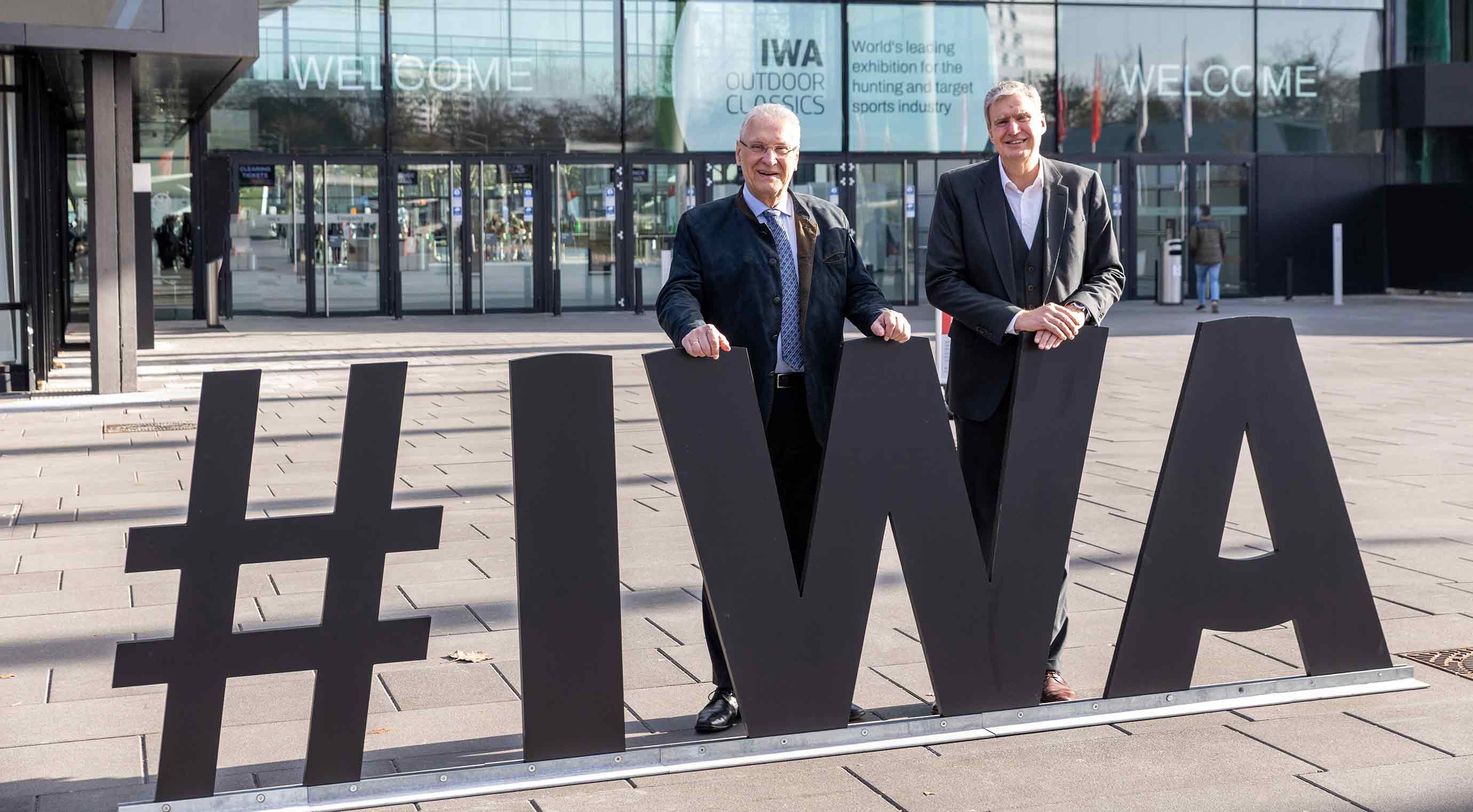 Joachim Herrmann and Peter Ottmann standing in front of the entrance to the IWA next to large black #IWA letters. The entrance area with the Welcome sign and trade fair graphics can be seen in the background.