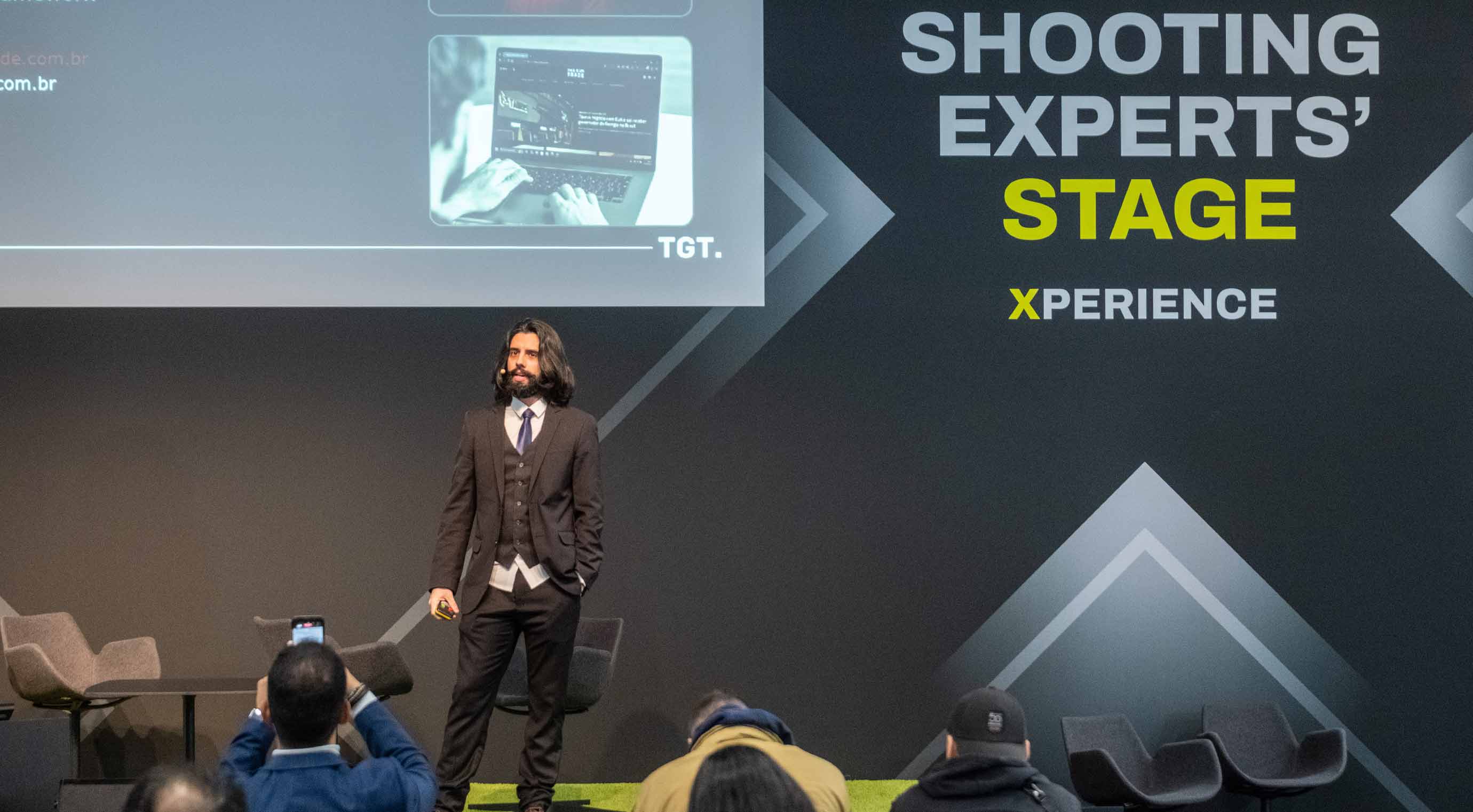 A person in a suit stands on stage in front of a large wall graphic with the words “Shooting Experts Stage,” while the audience listens in the foreground.