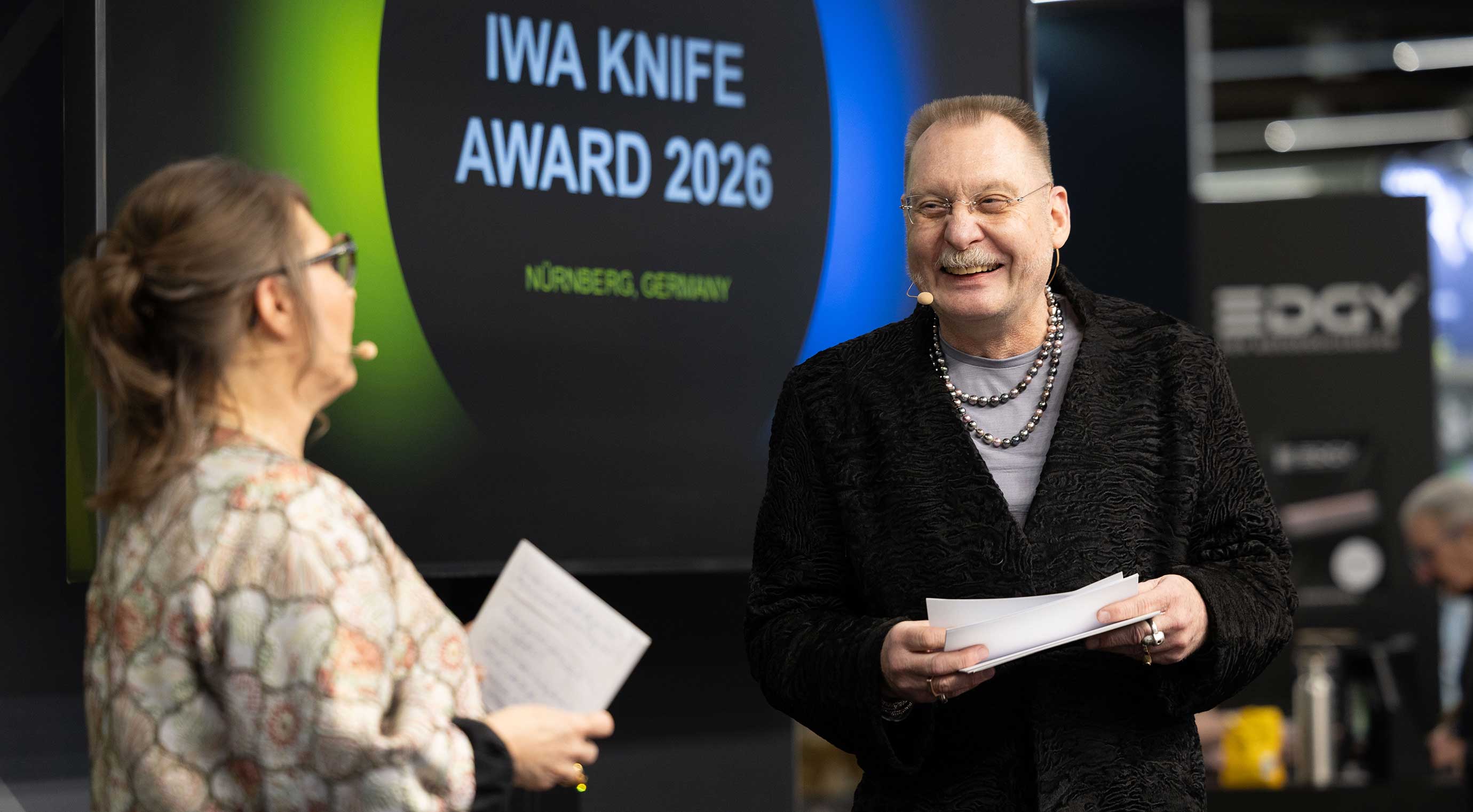 Two people are standing on a stage. In the background, there is a large screen with the words “IWA Knife Award 2026 – Nuremberg, Germany.”